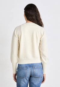 Cremfarbener, gestrickter Pullover mit Rippstruktur und Ballonärmeln. Er hat eine taillierte Taille und einen runden Ausschnitt, kombiniert mit blauen Jeans.