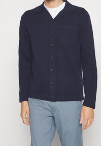 Marinblå cardigan i stickad tyg med kentrada krage, sju knappar och bröstficka. Långärmad med rak skärning.