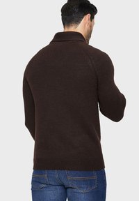 Brun strikket sweater med høj krave, ribbet tekstur og tætsiddende pasform, parret med blå denim jeans. Bagvinklen viser minimal detaljering.