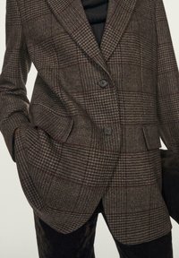 Blazer a quadri marrone realizzato in tessuto testurizzato con una vestibilità strutturata, chiusura a due bottoni e due tasche frontali. Sottotoni rossi sottili nel motivo.