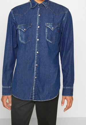 Chemise en denim bleu foncé à manches longues, avec un empiècement pointu, deux poches poitrine et une fermeture à boutons. Elle présente des coutures contrastantes et un ourlet arrondi.