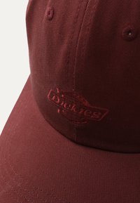 Bordeaux lue laget av teksturert stoff, med et preget logo. Designet har en buet skjerm og søm langs kantene.