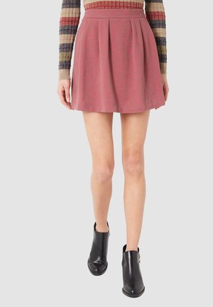 Mujer vestida con una falda corta plisada de color rosa oscuro, una blusa de rayas de manga larga y botas negras hasta el tobillo, de pie contra un fondo liso.