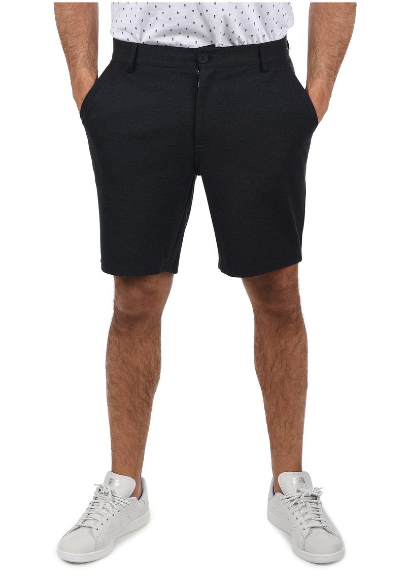 Pantalones cortos negros de una tela suave, con frente plano, bolsillos laterales y cierre de botón. Llevados con zapatillas gris claro.