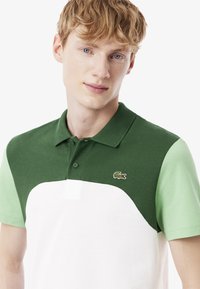 Vihreä ja valkoinen polo-paita, joka on valmistettu teksturoidusta kankaasta. Paita sisältää kahden sävyn designin, kolmen napin napituksen ja pienen logon rintapielessä.