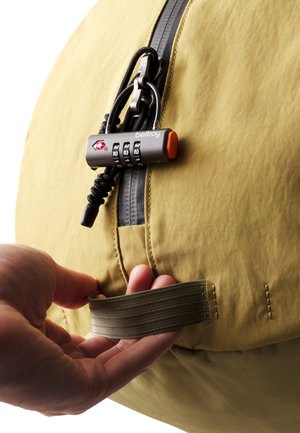 Mano che tiene la tracolla di una borsa giallo senape chiusa con un lucchetto a combinazione nero sul cursore della cerniera.