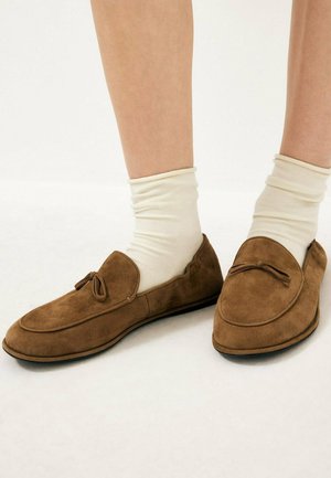 Lichtbeige sokken gedragen met bruine suède loafers met een klein gebonden strikdetail en platte zwarte zolen op een witte achtergrond.
