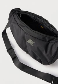 Μαύρη υφασμάτινη τσάντα χιαστί με ρυθμιζόμενο ιμάντα ώμου και μερικώς ανοιχτό φερμουάρ στην κύρια θήκη που δείχνει το άδειο εσωτερικό.