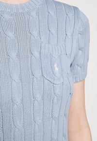 Jasnoniebieski sweter na drutach z wzorem warkoczowym, krótki rękaw oraz kieszonka na piersi z małym emblematem. Miękka faktura i dopasowany krój.