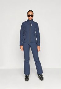 Marineblauer Skianzug und Hose aus strapazierfähigem Stoff, mit hohem Kragen, Reißverschlusstaschen und schlankem Schnitt. Glänzend schwarze Skischuhe vervollständigen den Look.