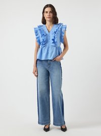 Kvinne iført en blå volangermed løs ermer topp med knappedetaljer og brede to-fargede blå jeans, står med en hånd i lommen.