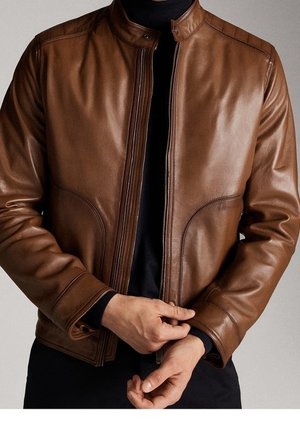 Mężczyzna w brązowej skórzanej kurtce i czarnym golfie zapinający zamek kurtki obiema rękami.