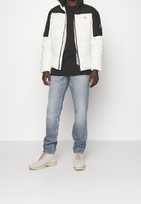 Mężczyzna w czarno-białej kurtce puchowej, czarnej koszulce, jasnoniebieskich dżinsach i beżowych zamszowych butach stoi na tle jednolitego tła.