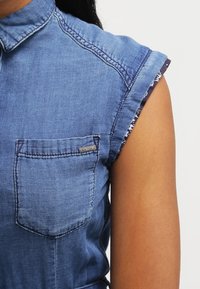 Koszula dżinsowa z kołnierzykiem, krótkimi, podwiniętymi rękawami, kieszenią na piersi oraz metką marki. Jasnoniebieski kolor z miękką teksturą i casualowym wzornictwem.