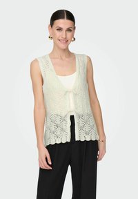 Gebreide mouwloze vest in licht mintgroen, met een gestructureerd kantpatroon, golfzoom en een sluiting met een strik aan de voorkant. Ggedragen over een witte top.