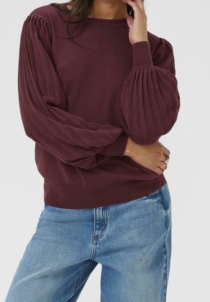 Femme portant un pull côtelé bordeaux avec des manches bouffantes et un jean bleu clair, main touchant le visage et la taille.