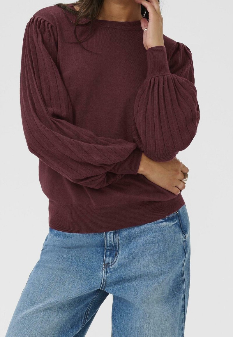 Femme portant un pull côtelé bordeaux avec des manches bouffantes et un jean bleu clair, main touchant le visage et la taille.