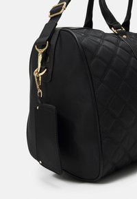 Borsa a tracolla nera trapuntata con forma arrotondata, hardware dorato, doppi manici superiori e un'etichetta removibile attaccata con un gancio metallico.