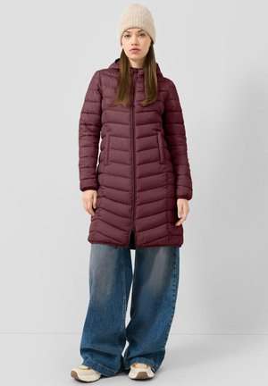 Femme portant une longue veste matelassée bordeaux, un jean bleu à jambes larges, des baskets blanches et un bonnet beige en tricot, debout devant un fond uni.