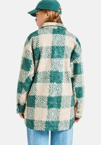 Een groen en crème geruite fleece jas met een kraag, oversized pasvorm, textuurstof en knoopdetails op de mouwen. Draagt met denim jeans.