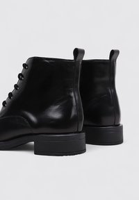 Bottes noires en cuir au niveau de la cheville avec une texture lisse, un bout rond et un petit talon empilé. Présente un laçage à l'avant et des languettes sur les côtés.