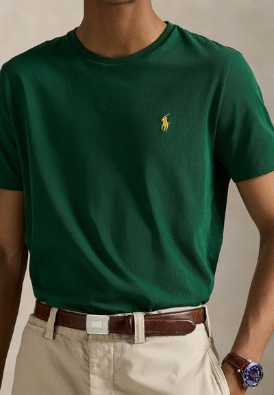 Πράσινο βαμβακερό μπλουζάκι  με στρογγυλή λαιμόκοψη, που διαθέτει ένα μικρό κίτρινο κεντημένο λογότυπο στο στήθος, συνοδευόμενο από μπεζ παντελόνι και καφέ ζώνη.