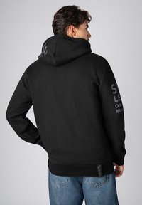 Zwarte hoodie met een gestructureerde afwerking, voorzien van een grote capuchon, zijkant branding en een aansluitende pasvorm. Gecombineerd met lichte spijkerbroek.