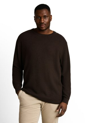 Homme portant un pull marron foncé et un pantalon beige, debout avec une main dans la poche devant un fond blanc uni.