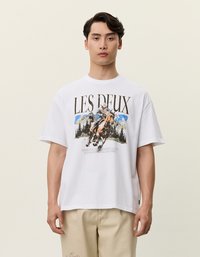 Biała bawełniana koszulka z grafiką przedstawiającą jeźdźca konnego, góry i drzewa; luźny krój, krótkie rękawy i okrągły dekolt.