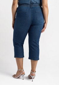Jeans recortados azul oscuro con una cintura elástica, que presentan dos bolsillos traseros y detalles plateados a lo largo de las costuras laterales. Combinados con tacones plateados.