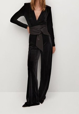 Kobieta ubrana w czarny aksamitny kombinezon z długim rękawem, głębokim dekoltem w serek i szerokim satynowym paskiem zawiązanym w talii, stojąca na tle jednolitej białej ściany.