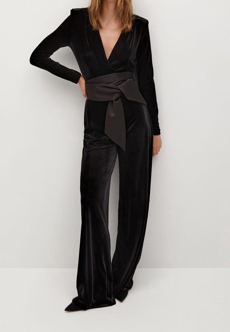 Femme portant une combinaison noire en velours à manches longues avec un décolleté en V profond et une large ceinture en satin nouée à la taille, posant devant un mur blanc uni.