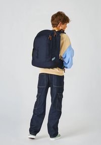 Sac à dos marine avec zips et accents marron, doté de bretelles rembourrées. Porté avec un pull à superpositions beige et bleu clair et un jean foncé.