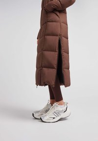 Abrigo de plumas marrón con diseño acolchado, cremallera lateral y cuello alto, combinado con zapatillas deportivas blancas y negras. Leggings visibles debajo.