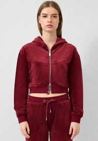 Burgunderfarbene, verkürzte Kapuzenjacke aus weichem Samt, mit einem Frontreißverschluss, Kängurutasche und gerippten Bündchen, sowie einem gestickten Logo auf der Brust.