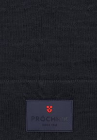 Ciemnoniebieska dziergana czapka beanie o miękkiej fakturze. Posiada prostokątną łatę z logo w czerwonych i białych akcentach, otoczoną niebieskim tłem.