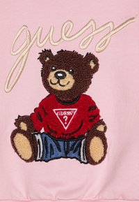 Felpa rosa ricamata con un orsetto che indossa un maglione rosso e jeans blu con dettagli neri e marroni. Logo "Guess" sopra l'orsetto.