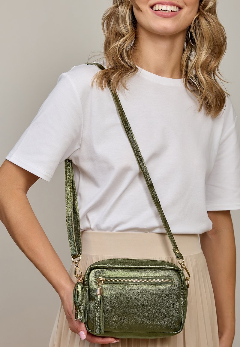 Žalias metalinis crossbody krepšys su auksinėmis detalėmis, su užtrauktuko kišene ir kutu, nešiojamas ant balto marškinėlio ir smėlio spalvos sijono.