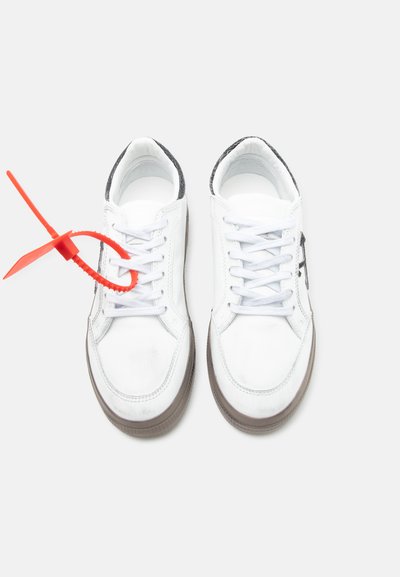 Ζευγάρι λευκών αθλητικών παπουτσιών με γκρι σόλες, ένα παπούτσι με ένα ευδιάκριτο κόκκινο πλαστικό ταμπελάκι προσαρτημένο στα κορδόνια, σε λευκό φόντο.