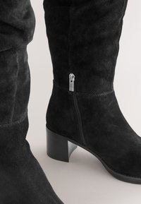 Schwarze Wildleder-Stiefel bis zur Mitte der Wade mit seitlichem Reißverschluss und Blockabsätzen auf neutralem Hintergrund, Nahaufnahme von Textur und Nähten.