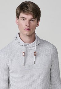 Lichtgrijze hoodie gemaakt van textuurgebreide stof. Voorzien van een kraag, trekkoorden met houten accenten en een subtiele gecamoufleerde logo op de voorkant.
