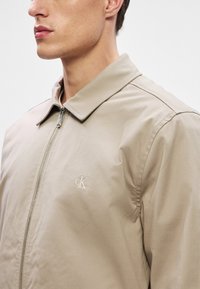 Beżowa, lekka kurtka z klasycznym kołnierzem, pełnym zamkiem błyskawicznym oraz wyszywanym logo po lewej stronie klatki piersiowej. Gładki, teksturowany materiał.