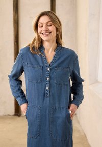 Femme souriante aux yeux fermés portant une robe en jean à manches longues avec une patte de boutonnage et quatre poches avant, les mains dans les poches à l'intérieur.