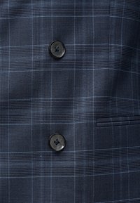 Ύφασμα κοστουμιού σκούρου μπλε με διακριτικό καρό σχέδιο, που διαθέτει δύο μαύρα κουμπιά και μια οριζόντια τσέπη με πλάγια ραφή.