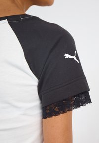 Tricou din bumbac alb și negru, cu mâneci scurte, decorat cu dantelă neagră la capete și cu un logo alb Puma pe mâneca stângă.