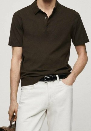 Mężczyzna w ciemno-brązowej koszulce polo z krótkim rękawem, w białych spodniach z czarnym paskiem, trzyma brązową torbę w lewej ręce, a prawą rękę ma w kieszeni.