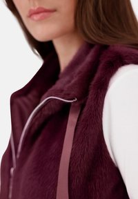 Burgunderfarbenes Kunstfellweste mit Reißverschluss und Satinband-Akzenten, kombiniert mit einem weißen Langarmshirt. Glatte Textur sichtbar.