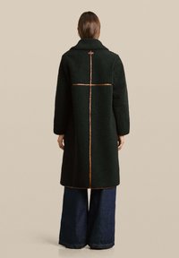 Persona con un lungo cappotto nero strutturato che presenta cuciture beige visibili a forma di croce sulla schiena, abbinato a pantaloni larghi di un blu scuro.