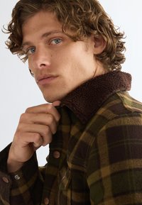 Jeune homme aux cheveux bouclés ajustant le col d'une veste à carreaux verte et marron avec un col texturé marron foncé.