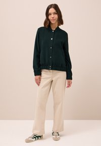 Donkergroene, knoopsluiting bomberjack met een losse pasvorm, elastische onderrand, ribgebreide manchetten en twee zijzakken, gedragen met beige broek.